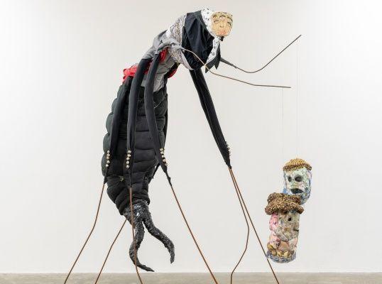 Image: sculpture of what looks like a very large black earwig with thin steel-bar-like legs and pretty terrifying pincers at the bum end; at the head end, we seem to see a pinned-on photo of a pale human face with too many eyes; the upper body seems to be wearing some sort of messed-up suit; a strange object appears to be attached to one of the critter’s legs – one or more heads made of strange material, papier-mâché or sweets or coloured cotton balls or…; this all seems to be in some sort of gallery space • “Curated by Sara Greavu ( Project Arts Centre) in 2023, Kian Benson Bailes’ Culchie boy, I love you / Grá mo chroí thú, mo chábóigín féin arrives at Mermaid Arts Centre in a new iteration. Comprising sculpture, digital & material collage, musical instruments, ceramics, textiles, woodwork, and live sculptural sound elements. Culchie boy, I love you / Grá mo chroí thú, mo chábóigín féin references Irish folklore, mythology, and craft traditions in an exuberant expression of rural…”