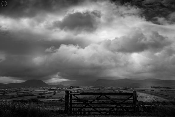 monochrome shot of gate and farmland with storm cloud field skies above and mountain range in distance