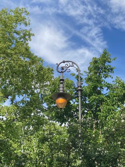 Alte Berliner Staßenlaterne, die auch am Tage brennt. Im Hintergrund grüne Bäume und blauer Himmel.