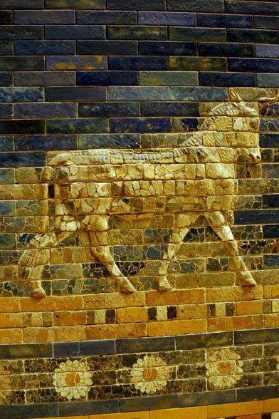 Ein Relief eines gehenden Tieres, möglicherweise eines Pferdes, das auf einem Mauerwerk aus bunten Ziegeln dargestellt ist. Die Ziegel sind in verschiedenen Farbtönen angeordnet, die eine strukturierte Wand bilden. Unter dem Tier sind florale Muster in Form von Blumen zu sehen.