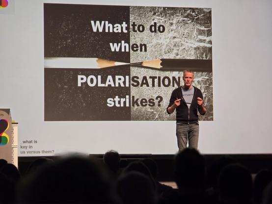 Ein Mann steht auf einer Bühne. Hinter ihm eine Präsentation mit dem Text What to do when Polarisation strikes