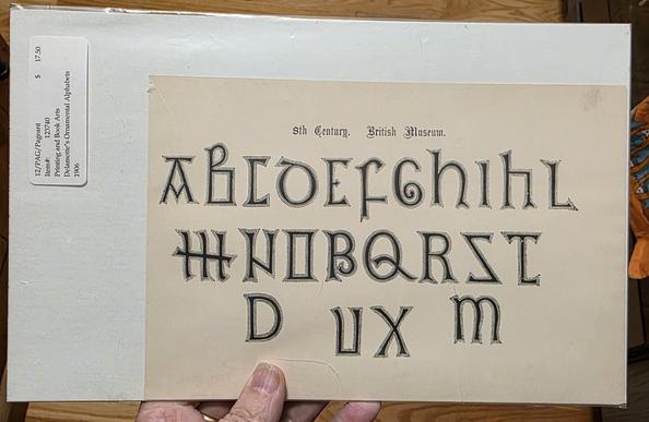 A photo of a hand holding a printed page  showing 23 ALLCAPS letters in 3 rows. Above them in blackletter small print is written '8th Century. British Museum. The letters have different line strokes from those popukar today. Missing are J, Z (the s looks like a reversed z) V (there is a u)