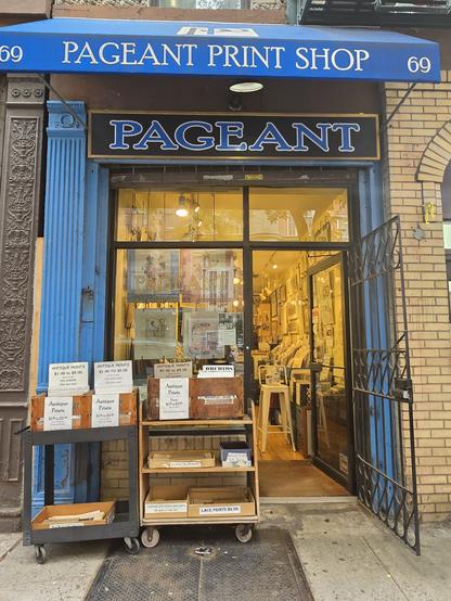 Photo of a shop calked 'Pageant' from with boxed on shekves on the street in frong. The boxed are full of prints. Inside we can see rows of boxes of printd