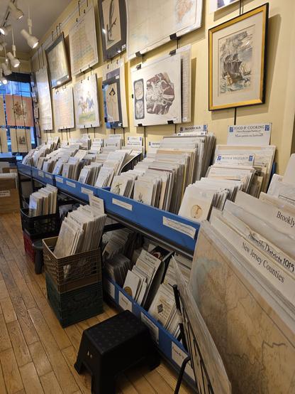 Photograoh of inside the store, lots of different size prints lined up like records in a vynil album record store. Buxes on thd floor, under the shelves and prints hung on the wall. A sense of crowdibg