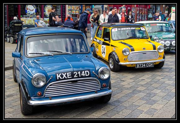 A street scene featuring several classic Mini cars parked on a cobblestone pavement. In the foreground, a blue Mini with the license plate "KXE 214D" is parked beside a yellow Mini with racing stripes and the number "34" on its side, with the license plate "B734 CGH." Further back, more classic Minis are visible, including a green one with the license plate "W46 DLY." People gather around the cars, chatting and enjoying the atmosphere. In the background, a café with outdoor seating hosts patrons, with signs advertising various food items. The setting suggests a car show or gathering of Mini enthusiasts.