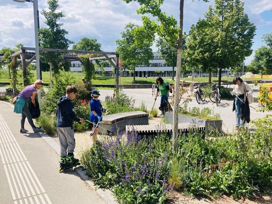 Das Bild zeigt die Seestadt Lounge und den Hannah-Arendt-Park dahinter! Man sieht zwei Erwachsene und zwei Kinder beim Müll sammeln. Der Himmel ist blau, mit einzelnen Wolken.
