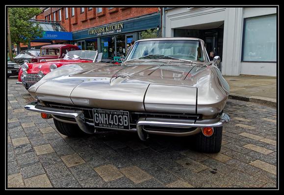 A classic 1964 Corvette Stingray in silver is parked on a cobblestone street in front of a restaurant named "Turquoise Kitchen." The Corvette's license plate reads "GNH403B" with the text "Retro Classic Car" below it. Behind the Corvette, several other vintage cars are lined up, including a red convertible. The setting appears to be an outdoor car show or gathering, with the cars beautifully maintained and arranged in front of shops and restaurants, creating a nostalgic atmosphere.
