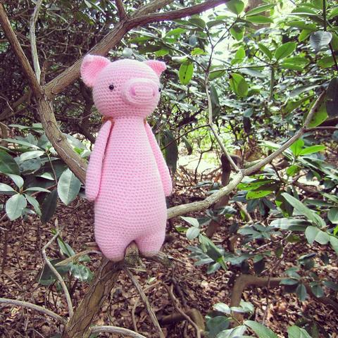 Picture of a crocheted amigurumi pig, standing upright in a tree, with the straps of a backpack visible on its shoulder. In the background are trees and bushes.
