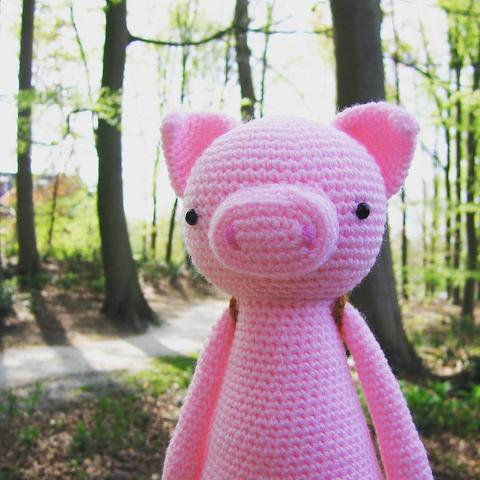 Portrait picture of a crocheted amigurumi pig, looking at the viewer. In the background is a path going through the trees. It's sunny.