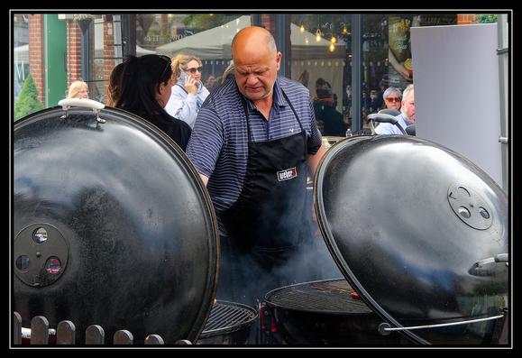 A man wearing a striped polo shirt and a black apron with the Weber logo is grilling food on two large charcoal grills. The grills have their lids open, revealing the cooking grates inside. Smoke rises from the grills, indicating active cooking. Several people stand around, some engaged in conversation. In the background, a brick building with a glass window is visible, along with a sign that reads "Perfect Coffee." The setting appears to be an outdoor event or gathering, with a communal atmosphere centered around cooking and socializing.