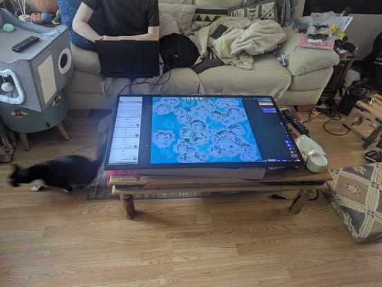 A large flat screen TV lies horizontally on a coffee table almost the same size as it, displaying terrain for an RPG battle. Behind it a tall goth enby sits crosslegged on a sofa, controlling the display on a laptop. A black cat slinks under the table and, while she's not actively doing anything suspicious in the photos I feel you can somehow tell from her posture that this cat lives to commit crimes.