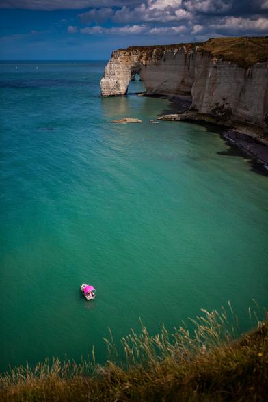 Coastal cliffs with a natural arch formation over turquoise waters, with a pink-roofed boat floating near the shore.