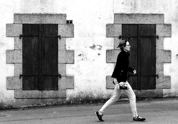 Black and white digital picture of a woman passing on the street. The woman is passing from left to right. Some motion-blur is visible. In the background Two windows with closed shutters can be seen.