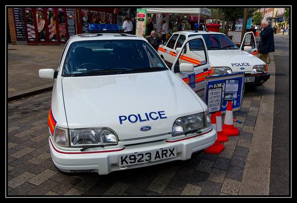 Two white police cars are parked on a paved street, both featuring blue emergency lights on top and the word "POLICE" written in bold blue letters across the front. The car in the foreground has the license plate "K923 ARX," while the one behind it has the license plate "LLYO." Both vehicles display red and yellow reflective stripes along their sides. Several orange traffic cones are positioned around the cars, indicating a designated area. To the right, an informational display board stands with various details. In the background, people walk and gather near a market stall adorned with colorful banners and signs. The scene suggests a public event or demonstration involving law enforcement.