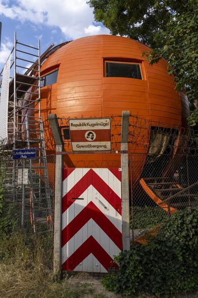 An orange sphere in a park with windows, surrounded by a fence and a gate. A sign reads "Republic Kugelmugel".