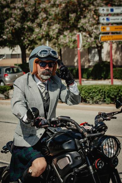 Urbano, medio plano desde 3/4 delantero-derecho. Voy conduciendo mi moto, una Yamaha XSR700 2019 modificada con la mano derecha y con la izquierda me llevo dos dedos a la visera del casco, un jet color gris medio, con forro color marrón claro y  antiparras sobre la visera.
Visto una chaqueta de príncipe de gales, un chaleco gris oscuro herringbone, una corbata de lana texturizada gris claro con rayas gris oscuro, un kilt black watch, guantes negros con la bandera de reino unido en el dorso de la mano y gafas oscuras de concha. En las medias, negras con borlas, asoma un puño de cuerno de ciervo. No hay cuchillo, solo el cuerno.
