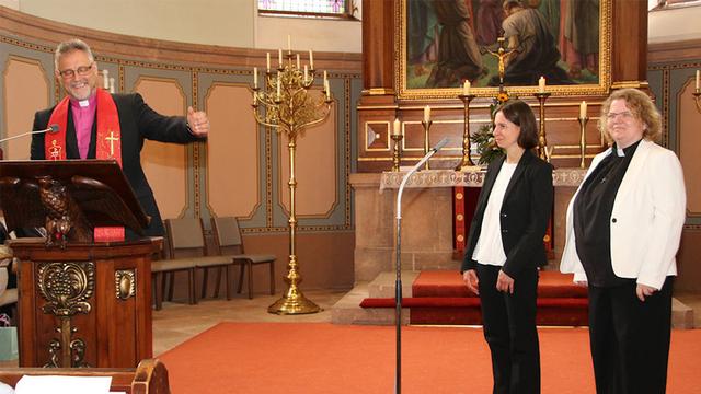 Im Chorraum einer älteren Kirche steht ein lächelnder Mann mit Kollarhemd, dunklem Jackett und Bischofsstola hinter einem Pult und weist mit ausladender Geste auf zwei junge Frauen hin, die etwa drei Meter entfernt stehen.