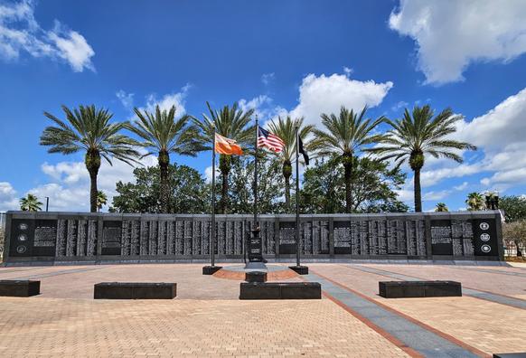 Large, black marble memorial marker, etched with the names of local veterans and their service, with a row of palms behind, and flags for the city of Jacksonville,  the state of Florida,  and the United States over an eternal flame.