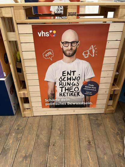 Ein Werbeplakat mit einem Mann mit Glatze und Brille, der ein weißes T-Shirt mit dem Text „Entschwörungstheoretiker“ trägt.