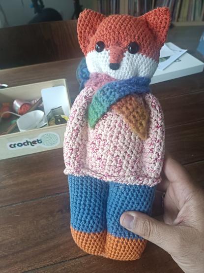 Doudou renard en crochet. Le doudou a le haut de la tête orange, le bas blanc, un pull en laine rose et blanc, une écharpe multicolore, un pantalon bleu et les pieds oranges.