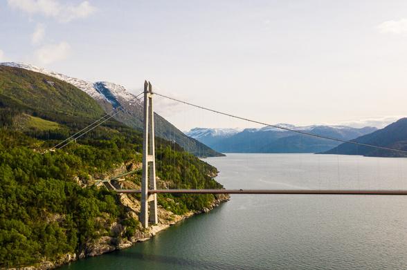 An aerial photo of a section of the Hardanger Bridge over the Hardanger Fjord against a backdrop of Norwegian mountains.