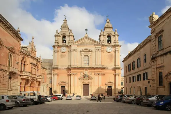 The image depicts a beautiful architectural scene featuring a church or cathedral, characterized by its ornate facade and two bell towers. The surrounding area appears to be a square with parked cars and buildings that likely reflect local architectural styles. The sky overhead is partly cloudy, contributing to the overall picturesque setting.

Image Credits: Wikimedia / Frank Vincentz / CC BY-SA 3.0