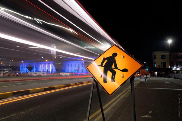 A nighttime city scene in Cork, Ireland, featuring a brightly lit orange roadworks sign with a silhouette of a person digging, set against a backdrop of light trails from passing vehicles and illuminated buildings. Visible text on a smaller sign in the background reads: "Batchelor's Quay ROAD CLOSED".