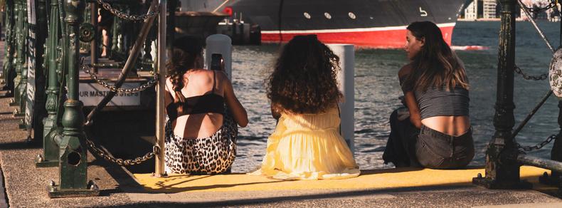 Three people sitting on a dock, facing the water. One person has a leopard-print skirt, one wears a yellow dress, and the third is in a striped top. A large ship with a red and black hull is in the background. Shadows are cast on the concrete surface, and metal railings run along the dock.