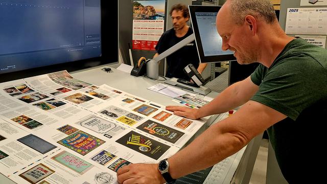 Man leaning over a table and smiling as he looks at a freshly printed sheet of colourful pages from a magazine about sign painting.