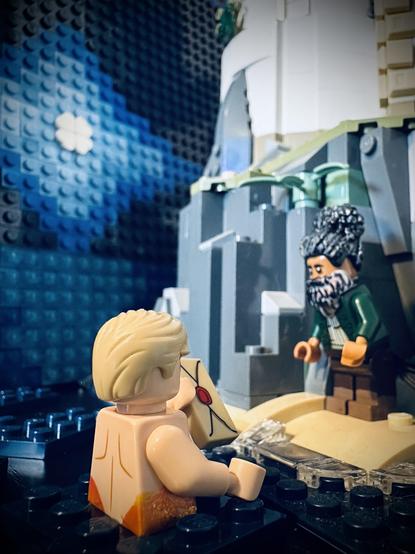 Lego MerStede rising out of the water and handing a letter towards Lego Ed, whose hair is up and has a full beard. He is standing on a beach below a lighthouse. The moon is full.