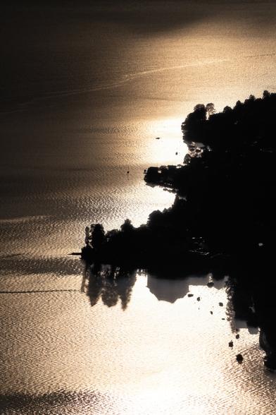 Gegenlichtaufnahme im Hochformat von einem Berg auf eine Uferpartie an einem See.