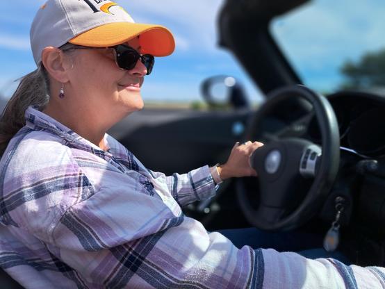 My hot wife having fun driving through twists and turns of the rural Indiana highway system. She wears a University of Tennessee ball cap, has a purple flannel shirt, I've hand on the steering wheel, and I've hand in the stick shift. The landscape in the distance is a blur behind her.