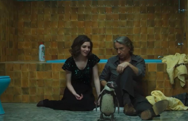 Een vrouw en man zitten op de grond in een badkamer met geel-bruine tegels, voor hen staat een pinguïn.