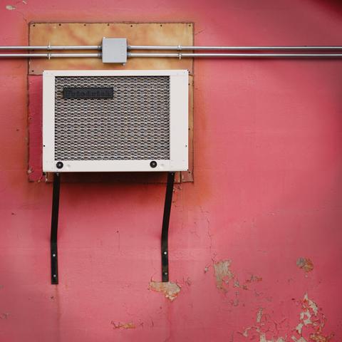 Wall-mounted air conditioning unit framed by exposed conduit and peeling pink paint. Centered composition with visible surface decay and saturated color contrast.