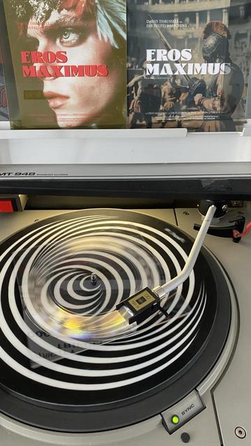 Durchsichtige, quadratische 7 Inch Vinyl Single auf einem Plattenspieler. Im Hintergrund die Hülle mit dem Titel „Eros Maximus“