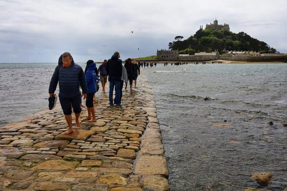 People walking on a causeway. The sea is partially covering the path. People are wading through the water. A small island with a castle is visible at the other end of the causeway.