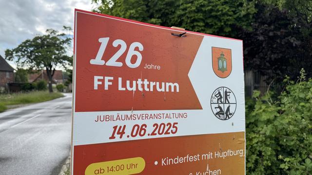 Wertetafel (in rot/weiß) für ein Jubiläum 126 Jahre der Feuerwehr in Luttrum im Juni 2025. Im Hintergrund Sträucher, Bäume, Dorfstraße und Gebäude.