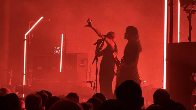 A stage is cloaked in smoke and red light. On the left, a man is standing with one hand raised and a microphone in the other hand. A fierce lady guitarist with long black hair stands next to him