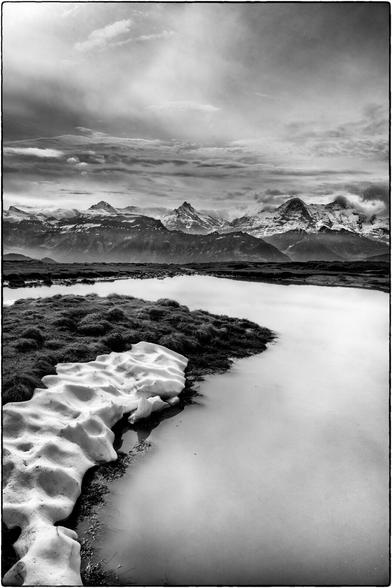 Schwarzweissfotografie im Hochformat eines Teichs mit Schneeresten am Rand. Im Hintergrund sind die Berner Alpen zu sehen.