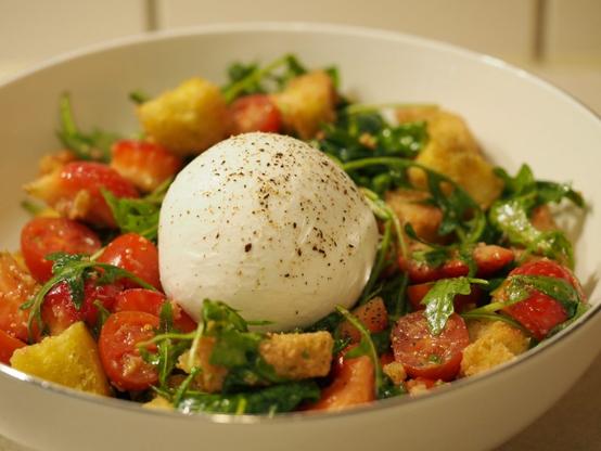 Eine große Portion des Salates in einer weißen Schüssel. In der Mitte liegt eine noch ganze Burrata Kugel, die mit gemahlenem Pfeffer bestreut wurde.