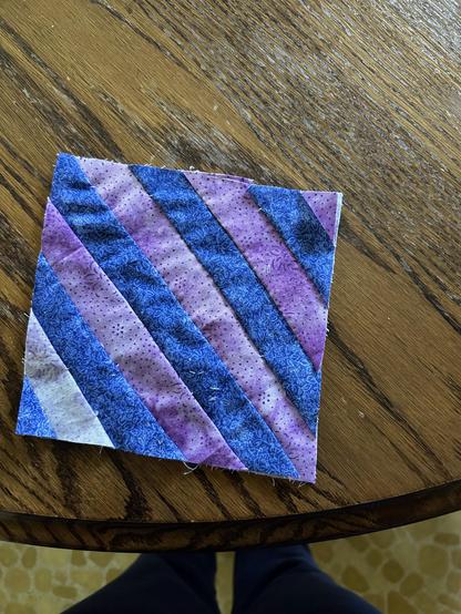 Quilt block with diagonal stripes. The block is a square piece of fabric featuring alternating diagonal strips of blue and purple fabrics. The blue strips have a subtle floral pattern, while the purple strips are lighter with small dotted designs. The block is placed on a wooden table, and the edge of the table is visible at the bottom of the image. Some legs and feet, likely of the person taking the photo, are faintly visible at the bottom edge. The block appears to be handmade, with visible stitching and slightly uneven edges.