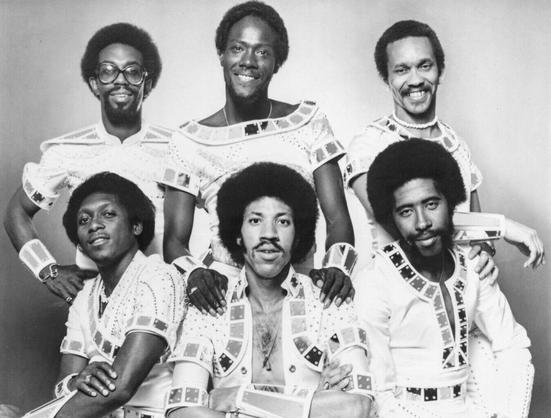 Commodores in 1970 in a Motown publicity shot, two years after their formation. They are wearing era-appropriate splangly, science fiction inspired outfits. Three members stand behind three seated members.