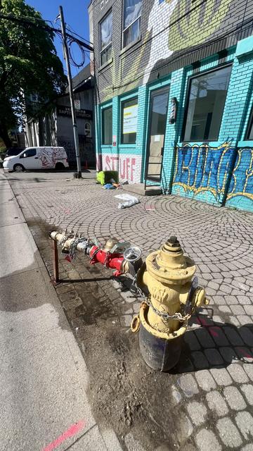 Yellow fire hydrant in the foreground has multiple attachments to one side, extending so far they are propped up by a piece of metal. There is water in the sidewalk. In the background is a closed shop, painted aqua with the word STUFF written on it in red. In front of it is it knocked over garbage bin with garbage on the ground. In the far background you see a white van half covered in graffiti and a baptist church. The more you look the odder it is.