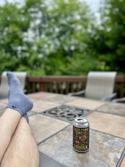 A pair of feet in gray socks rests on a table, alongside a can of "Oriole" beer from Beer Farm. The background features lush green foliage, and part of a wooden railing is visible.