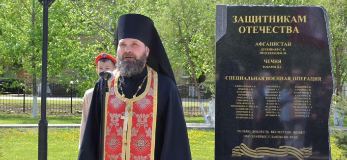 Denkmal mit Namen in kyrillischer Schrift, russisch-orthodoxer Priester daneben, Kind dahinter.
