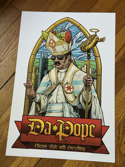 A photo of a print of Da Pope on a hardwood floor. The print itself is a  white man with a robust dark mustache wearing sunglasses along with the formal white and gold garb of the pope. The white cape has medallions on either lapel showing red 6-pointed stars as seen on the Chicago flag, and two sky blue bars wrap around the cape horizontally, also in reference to the flag. His belt/sash is also decorated with blue bars. The space inside his mitre is filled with oversized Chicago hot dog toppings (tomato, sport peppers, green relish, a pickle) and some yellow mustard is running down the front of the hat which also bears the Chicago municipal device, a Y in a circle, representing the Chicago River. In his left hand he’s holding a gold staff topped by a gilded Italian beef sandwich with a little halo and wings and he’s making a blessing gesture with his right hand. He's framed by a gothic arched window that contains a stained glass rendition of down Chicago looking past the top of the Sears Tower out toward the green lake and blue sky. At the bottom of all this is a red banner with large blackletter text that says 'Da * Pope' separated by a gold 6-pointed star, and below that in smaller blackletter text reads 'Chicago-style with Everything'.