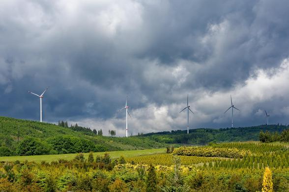 Auf einem Höhenzug sind fünf WEA vor düsteren Wolken zu sehen. Im Vordergrund Wiesen und Bäume.