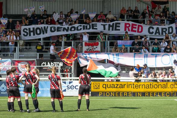 Clapton CFC players facing the stands @ Gallagher Stadium, where a large banner reading SHOW GENOCIDE THE RED CARD is displayed by fans.