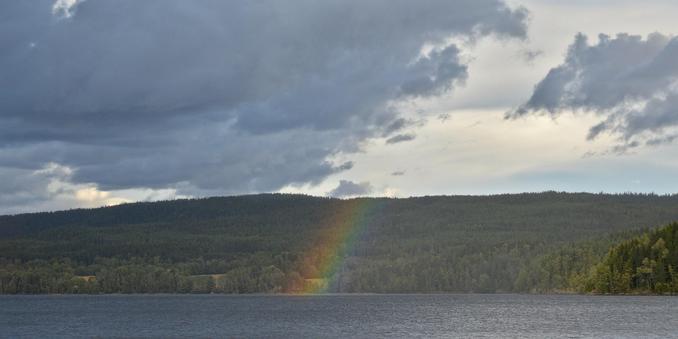 A photo of a lake with a forested hill on the far shore. There is a rainbow over the lake that is only visible against the hill. The sky is filled with light grey clouds with large dark clouds beneath them.