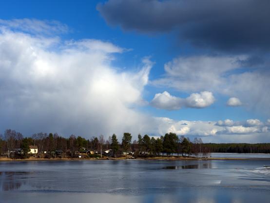 Calm lake reflecting a partly cloudy sky, with some dark clouds nearby to the right. Bare springtime trees can be seen on the peninsula in distance, with a few houses. The waters have some remaining sheets of ice.